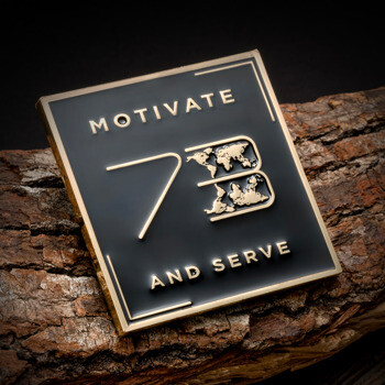 Square black enamel challenge coin featuring gold text Motivate 73 and Serve with a gold world map inlay on a wood bark background.