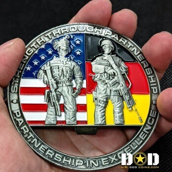 A challenge coin featuring two soldiers standing in front of the American and German flags with the text Strength Through Partnership.
