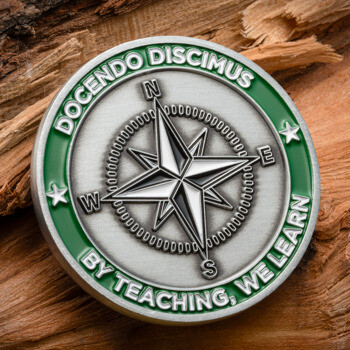 Antique silver challenge coin featuring a central compass rose, green enamel border with the text Docendo Discimus, and the phrase By Teaching, We Learn.