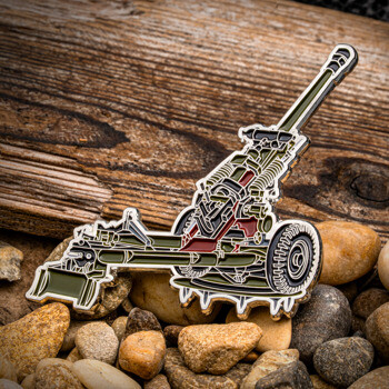 Custom enamel lapel pin shaped like a detailed military artillery howitzer cannon resting on small stones against a wooden background.