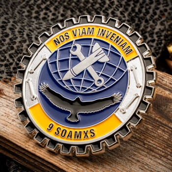 Gear-shaped challenge coin featuring 9 SOAMXS text, a bird silhouette, and a crossed wrench and bomb emblem on a blue and yellow background.