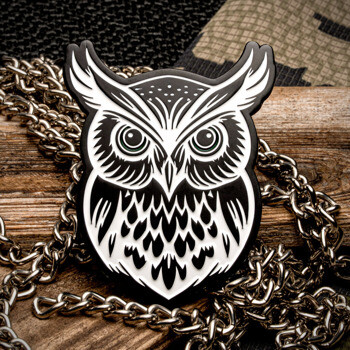 Black and white stylized owl lapel pin displayed on a piece of wood with a silver chain and camouflage fabric background.