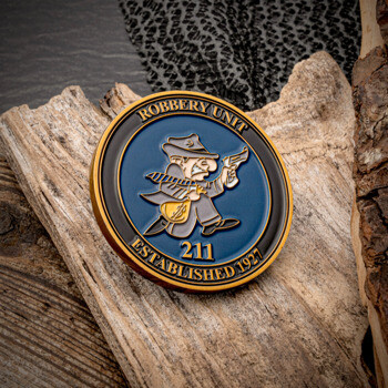 Circular robbery unit challenge coin featuring a cartoon detective character with a gold bag and pistol on a blue background, resting on wood bark.
