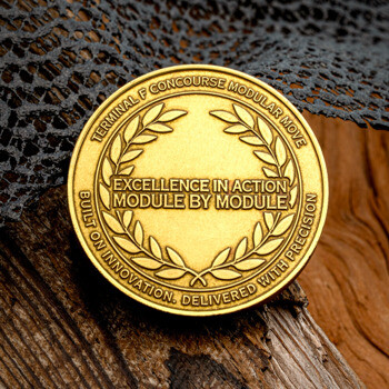 Gold challenge coin with laurel wreath and Excellence in Action Module by Module inscription on a wood background.