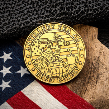 Gold challenge coin for Artillery Tea Co. featuring a skull in tactical gear holding a mug, set against an American flag and wood background.