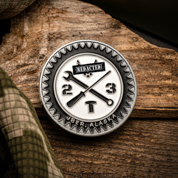 Circular challenge coin with crossed tools, a gear border, and text reading JBER Alaska 23T on a wood and camo background.