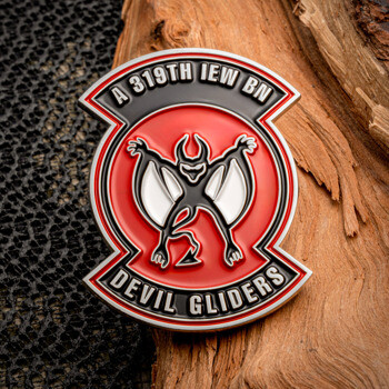 A 319th IEW BN Devil Gliders challenge coin featuring a red and black circular emblem with a devil figure on a piece of wood.