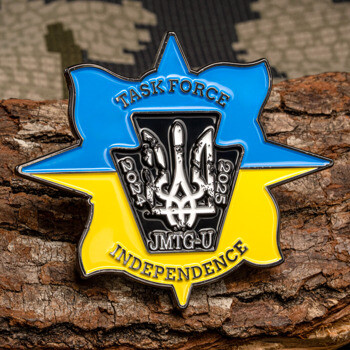 Blue and yellow shield lapel pin featuring Task Force Independence JMTG-U text and a central crest resting on a piece of tree bark.