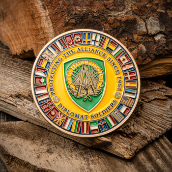 Gold challenge coin featuring a ring of international flags, a green shield emblem, and 'Diplomat Soldiers' text resting on wood.