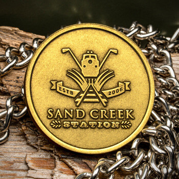Gold Sand Creek Station challenge coin featuring a train and golf club logo resting on a silver chain and wood.