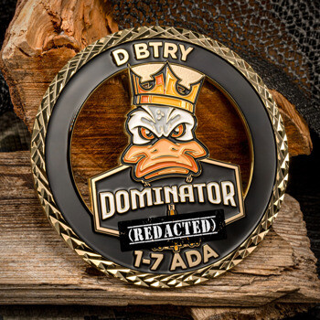 Gold challenge coin with a cut-out design featuring a crowned duck mascot and the text D BTRY Dominator 7-7 ADA against a black background.
