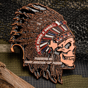 Skull in a Native American headdress challenge coin with red, white, and blue accents on a camouflage background.