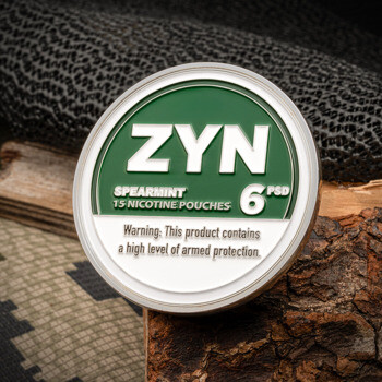 Circular challenge coin designed to look like a green and white ZYN spearmint nicotine pouch can, resting on a log with camo fabric.