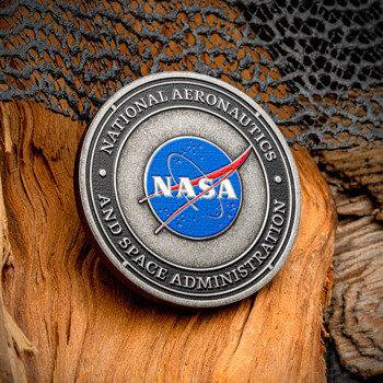 Antiqued silver NASA challenge coin with blue 'meatball' logo on a wood background.
