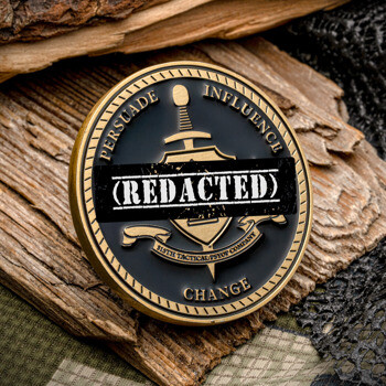Antique gold challenge coin with black enamel, featuring a sword and shield design with (REDACTED) text, set on wood and camouflage fabric.