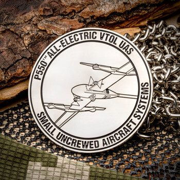 Silver challenge coin featuring a P550 All-Electric VTOL UAS aircraft design, resting on a wooden log and camouflage fabric.