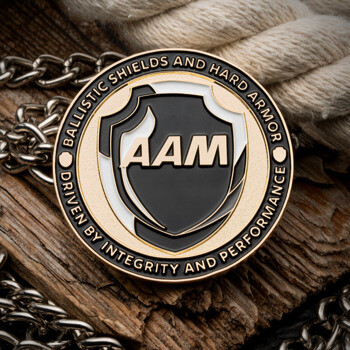 A round AAM challenge coin with a black shield logo and gold text, displayed on a wooden surface with a chain and rope.