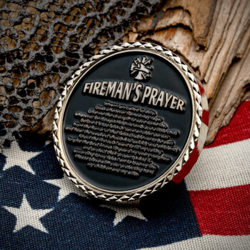 A round challenge coin featuring the Fireman's Prayer and a diamond-cut edge, resting on an American flag.