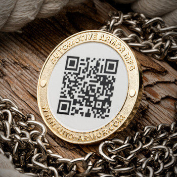 Gold challenge coin featuring a black and white QR code, the text Automotive Armor MFG, and a website URL, displayed on wood with a metal chain.