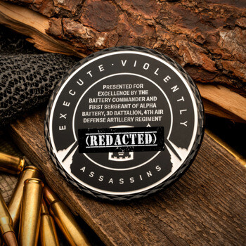 Black and white challenge coin for Alpha Battery, 3d Battalion, 4th Air Defense Artillery Regiment, displayed with rifle ammunition and wood.