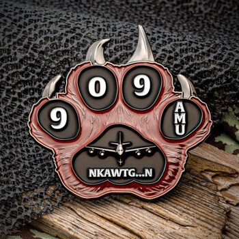 Paw-shaped challenge coin featuring numbers 9, 0, 9, and AMU text with a military aircraft silhouette on a textured red and black background.