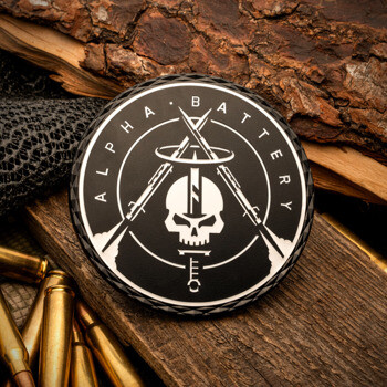 Black and silver Alpha Battery challenge coin featuring a skull and crossed rifles, displayed with ammunition and wood texture.