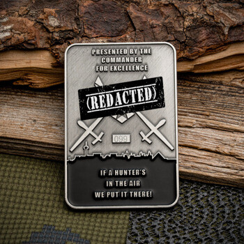 Rectangular silver challenge coin with 'REDACTED' stamp, drone silhouettes, city skyline, and military text over a wood and camouflage background.