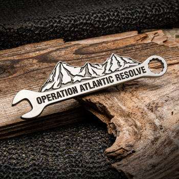 Silver wrench-shaped lapel pin featuring mountain peaks and Operation Atlantic Resolve text on wood background.