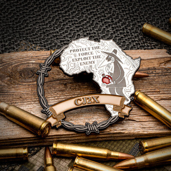 Custom lapel pin featuring the continent of Africa with a Spy vs Spy character, barbed wire, and CJ2X text, displayed among rifle cartridges.