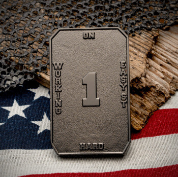 Rectangular antiqued metal challenge coin with the number 1 and text Working Hard On 1 Easy St, displayed over an American flag and netting.
