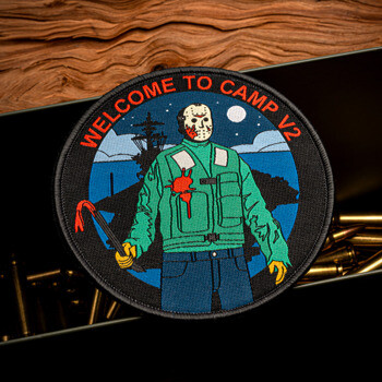 Circular embroidered patch featuring a masked character with a crowbar and the text Welcome to Camp V2, displayed on an ammunition box.