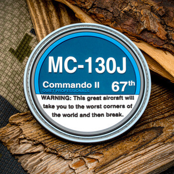 Circular challenge coin featuring MC-130J Commando II 67th text and a warning message, resting on tree bark and camouflage fabric.