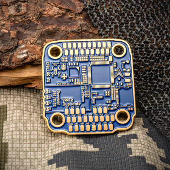 Square circuit board design challenge coin with blue enamel and gold recessed technical details, shown on camouflage fabric.