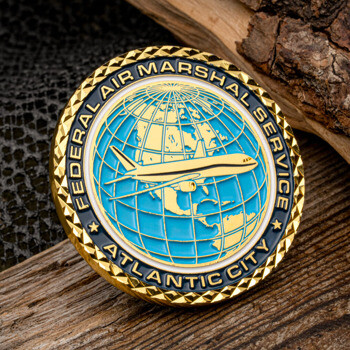 Gold diamond-cut edge challenge coin featuring the Federal Air Marshal Service logo with a plane over a blue globe, resting on wood and mesh.