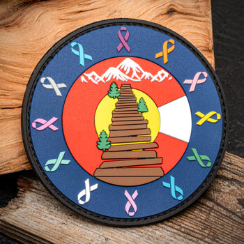 Circular PVC patch featuring a mountain landscape, wooden stairs, and colorful awareness ribbons around the border.