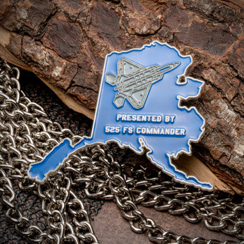 Alaska shaped challenge coin in blue enamel featuring an F-22 Raptor jet and 525 FS Commander text, displayed on tree bark with silver chains.