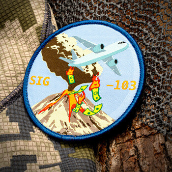 Circular embroidered patch featuring a white cargo plane flying over an erupting volcano with burning money and a blue border.