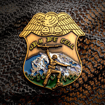 Gold shield-shaped challenge coin featuring an eagle, a soldier holding a rifle aloft, and a mountain backdrop with JBLM DFE CELL text.