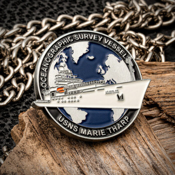 A circular USNS Marie Tharp challenge coin featuring an oceanographic survey vessel over a blue and silver world map, resting on wood and a chain.