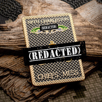 Rectangular gold challenge coin with diamond plate texture and NPTU Charleston Chiefs Mess text, resting on weathered wood.