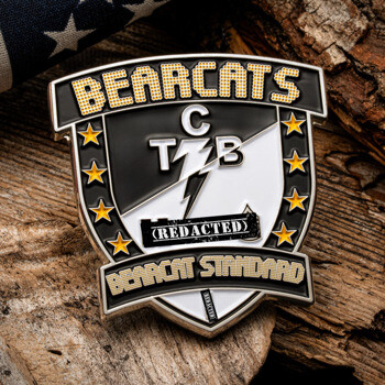 Shield-shaped lapel pin with Bearcats text, gold stars, and a lightning bolt design on a wood background with an American flag.