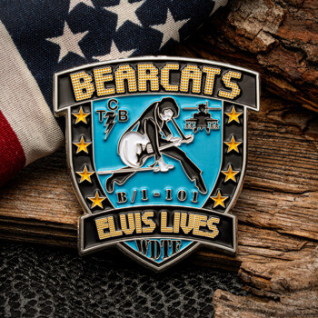 Shield-shaped challenge coin featuring an Elvis Presley silhouette, stars, and the text Bearcats B/1-101 Elvis Lives resting on wood next to an American flag.