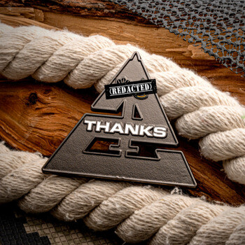 Triangular black and silver lapel pin with the word THANKS and a redacted text bar, displayed with rope and wood.