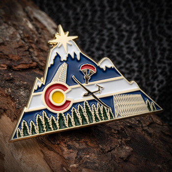 Mountain-shaped lapel pin featuring the Colorado flag emblem, a glider, parachute, pine trees, and a shining star at the peak.