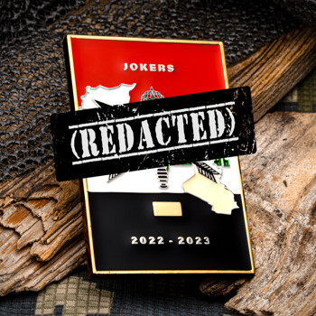 Rectangular Jokers 2022 to 2023 challenge coin with red, white, and black enamel details, resting on weathered wood and camouflage mesh fabric.