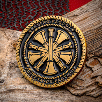 Gold and black fire chief challenge coin with a central speaker bugle symbol, inscribed with Serving Proudly and Chief Jason Parthun, displayed on wood.