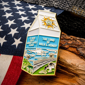 Milk carton shaped lapel pin featuring St. Johns County Sheriff's Office insignia, a road scene with bees, and text reading The Land of Milk & Honey.