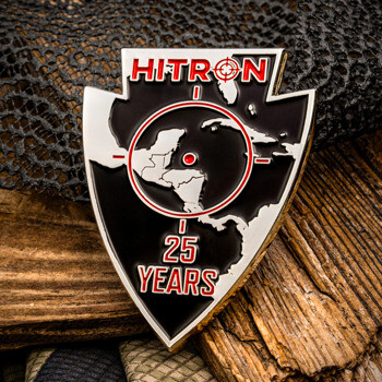 Arrowhead-shaped HITRON 25 Years lapel pin with a black enamel background, a map of the Caribbean, and a red crosshair design over a wooden background.