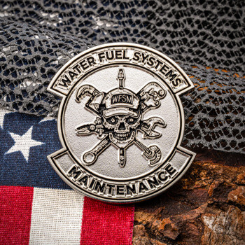 Silver challenge coin for Water Fuel Systems Maintenance featuring a skull and crossed wrenches design, resting on an American flag and rustic wood.