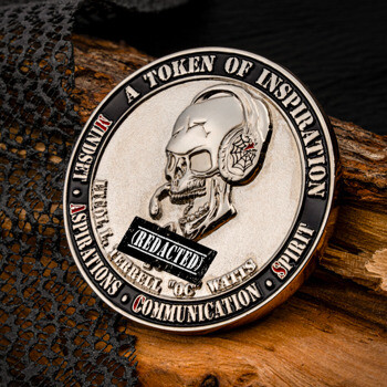 A round silver and black challenge coin featuring a skull wearing headphones with the text A Token of Inspiration, resting on a piece of wood.